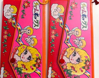 May include: Two rectangular plastic pencil cases in bright pink and red with orange trim. Each case features a cartoon illustration of a blonde character, small cherubs, flowers, and Japanese text. A yellow cord is attached to one, a red to the other.