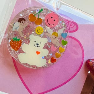 May include: A clear, round phone grip with a glittery interior. The grip features a white teddy bear, cherubs, cherries, a strawberry, and smiley faces. Colorful hearts and glitter are scattered throughout the design. The grip is on a pink heart-shaped background.