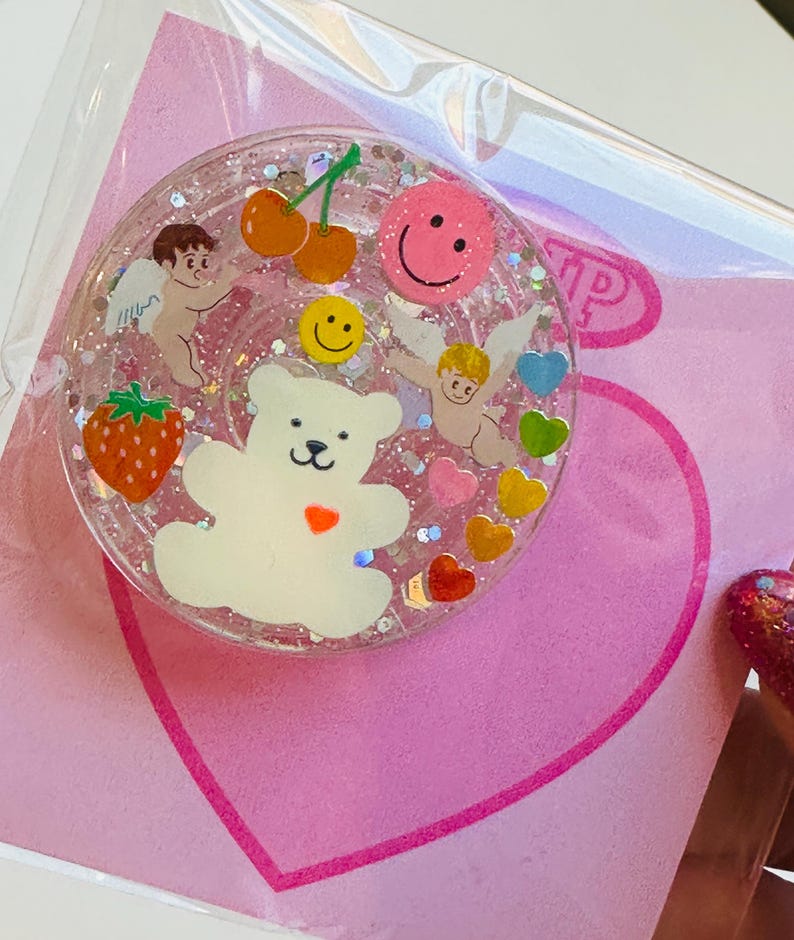 May include: A clear, round phone grip with a glittery interior. The grip features a white teddy bear, cherubs, cherries, a strawberry, and smiley faces. Colorful hearts and glitter are scattered throughout the design. The grip is on a pink heart-shaped background.