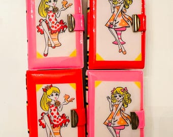 May include: Four vintage, rectangular, pink and red cases with a cartoon girl illustration. Each case has a gold-coloured clasp and a pink or red tab closure. The illustrations feature a blonde girl in a dress.