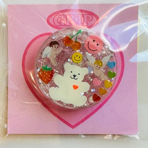 May include: A clear, round phone grip with a glittery interior, featuring a white bear, cherubs, cherries, a strawberry, and smiley faces. The grip is on a pink heart-shaped backing with the word "GRIP" in a pink oval.