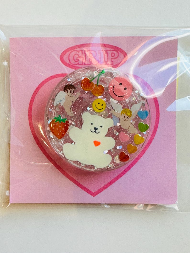 May include: A clear, round phone grip with a glittery interior, featuring a white bear, cherubs, cherries, a strawberry, and smiley faces. The grip is on a pink heart-shaped backing with the word "GRIP" in a pink oval.