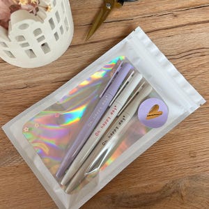 Könnte beinhalten: Eine transparente Plastiktüte mit drei Stiften in Pastellfarben: Lavendel, Weiß und Beige. Jeder Stift ist mit dem Schriftzug "Oh happy day" bedruckt. Ein Aufkleber mit einem herzförmigen Design ist ebenfalls sichtbar. Die Tüte liegt auf einer Holzoberfläche.