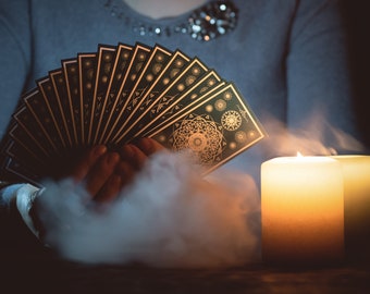 May include: A person holds a spread of tarot cards in front of a lit candle, with smoke rising from the candle. The cards have intricate gold designs on a dark green background.