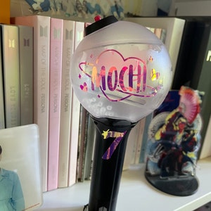 Peut inclure: Un b&acirc;ton lumineux en plastique transparent avec une poign&eacute;e noire et un c&oelig;ur rose avec le mot "Mochi" &agrave; l'int&eacute;rieur. Le b&acirc;ton a un design argent&eacute; paillet&eacute; et une sangle noire.