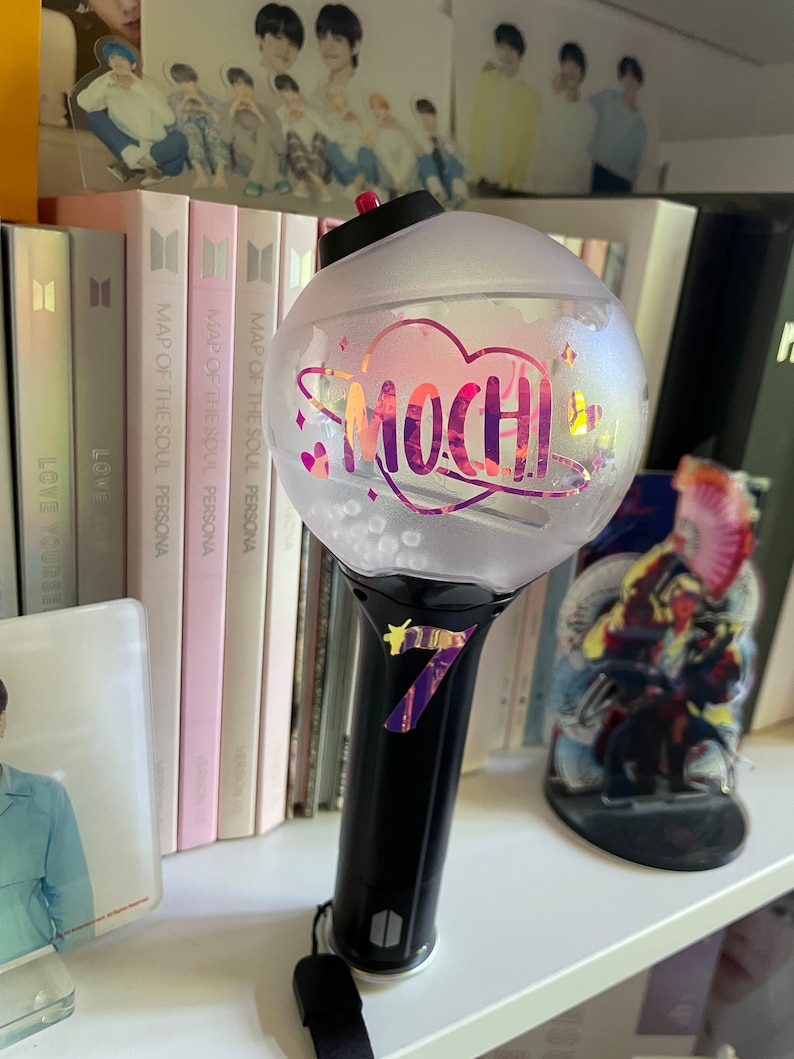 Peut inclure: Un b&acirc;ton lumineux en plastique transparent avec une poign&eacute;e noire et un c&oelig;ur rose avec le mot "Mochi" &agrave; l'int&eacute;rieur. Le b&acirc;ton a un design argent&eacute; paillet&eacute; et une sangle noire.