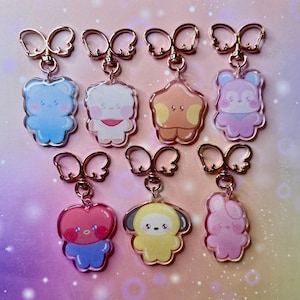 Könnte beinhalten: Eine Sammlung von neun Acryl-Schlüsselanhängern mit Cartoon-Charakter-Designs. Jeder Schlüsselanhänger zeigt einen anderen Charakter in verschiedenen Farben, darunter Blau, Weiß, Orange, Rosa und Gelb. Die Schlüsselanhänger haben goldfarbene, bogenförmige Verschlüsse.