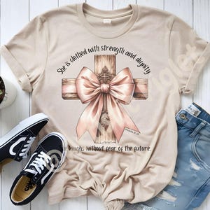 May include: A beige t-shirt featuring a cross adorned with a pink bow. The text "She is clothed with strength and dignity" and "Proverbs 3:35" is printed on the shirt. The phrase "laughs without fear of the future" is also printed on the shirt.