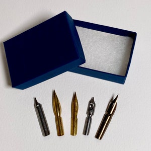 May include: A blue box with a white interior contains five different nibs for calligraphy pens. The nibs are made of metal and have a variety of shapes and sizes.