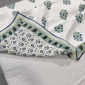 Può includere: Un copriletto trapuntato bianco con un motivo floreale ripetuto in blu e verde. L'angolo ripiegato rivela un bordo a contrasto con una fitta stampa di viticci e fiori blu e verdi, rifinito con un profilo a cordoncino blu scuro.