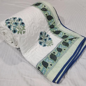 Può includere: Una coperta trapuntata bianca con un motivo floreale ripetuto stampato in blu e verde. La coperta presenta un bordo largo con un design a righe verdi e motivi floreali blu scuro, rifinito con un bordo in tessuto blu scuro.