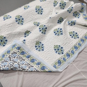 Può includere: Una coperta trapuntata bianca con un motivo ripetuto di piccoli grappoli di fiori blu con foglie verdi. I bordi sono rifiniti con una bordura decorativa che presenta una fitta stampa di tralci floreali nei toni del blu e del verde.