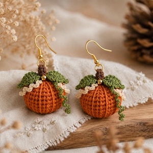 Può includere: Un paio di orecchini pendenti a forma di zucca all'uncinetto color arancione, con piccioli marroni, foglie verdi e una piccola perlina dorata. Ogni zucca è decorata con un bordo ondulato color crema e un viticcio verde pendente.