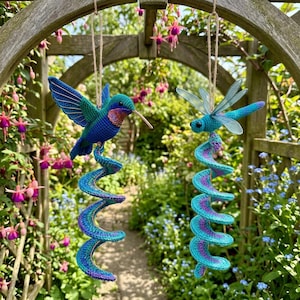 Op de afbeelding: Twee hangende gehaakte ornamenten met een blauwe en groene kolibrie en een blauwe libel, elk bevestigd aan een spiraalvormige, blauw en paars gekleurde staart. Beide figuren hangen aan dunne touwtjes voor weergave.
