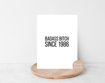 May include: A white card with the text "BADASS BITCH SINCE 1986" in black. The card rests on a wooden tray, with a small sticker reading "Blank Inside". A simple, bold design.