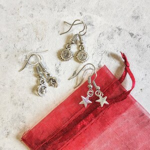 May include: A set of silver earrings featuring a sun, moon and star design. The earrings are presented in a red organza bag.