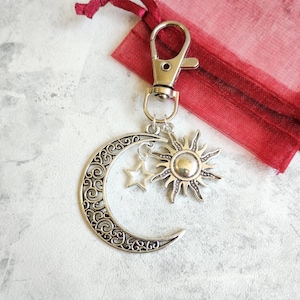 May include: A silver-toned metal keychain featuring a crescent moon charm with intricate scrollwork, a small five-pointed star charm, and a sun charm with radiating rays. The charms are attached to a silver-toned lobster claw clasp.