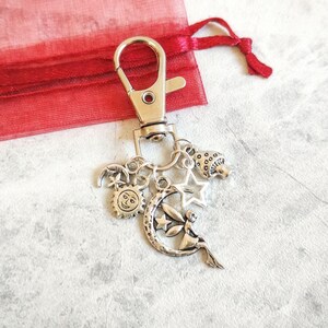 May include: A silver-toned metal keyring featuring a lobster clasp and multiple dangling charms. The charms include a sun with a face, a crescent moon with a fairy, a five-pointed star, and a small mushroom, all attached by jump rings.