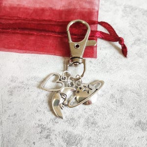 May include: A silver-toned metal keychain featuring a lobster clasp attached to three charms: a heart outline, a crescent moon with a face, and a planet with a ring and a star. The charms are suspended from a circular metal ring.