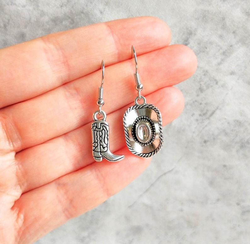 May include: A pair of silver dangle earrings with a cowboy boot charm on one earring and a cowboy hat charm on the other earring.