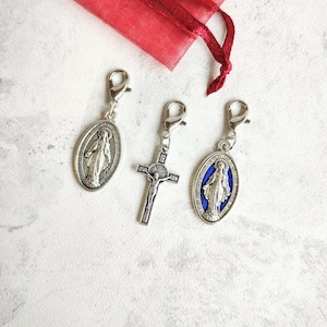 Könnte beinhalten: Drei religiöse Anhänger mit silberfarbenen Verschlüssen. Zwei ovale Anhänger zeigen eine religiöse Figur, und einer ist ein Kruzifix. Ein roter Stoffbeutel befindet sich im Hintergrund.