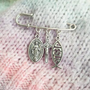 May include: A silver safety pin featuring three religious charms. These include an oval medal with a figure, a crucifix, and a fish-shaped pendant with the word "JESUS". The pin is set against a pink and purple knitted background.