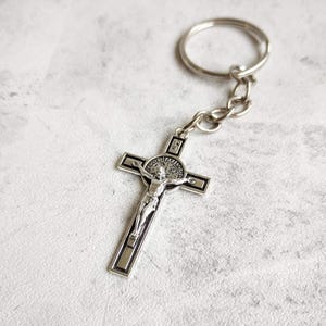 May include: Silver crucifix keyring. The cross has a detailed depiction of Jesus Christ and is attached to a chain and a ring. The design includes a detailed figure of Jesus on the cross, with the words "PAX" and "VRS" visible.
