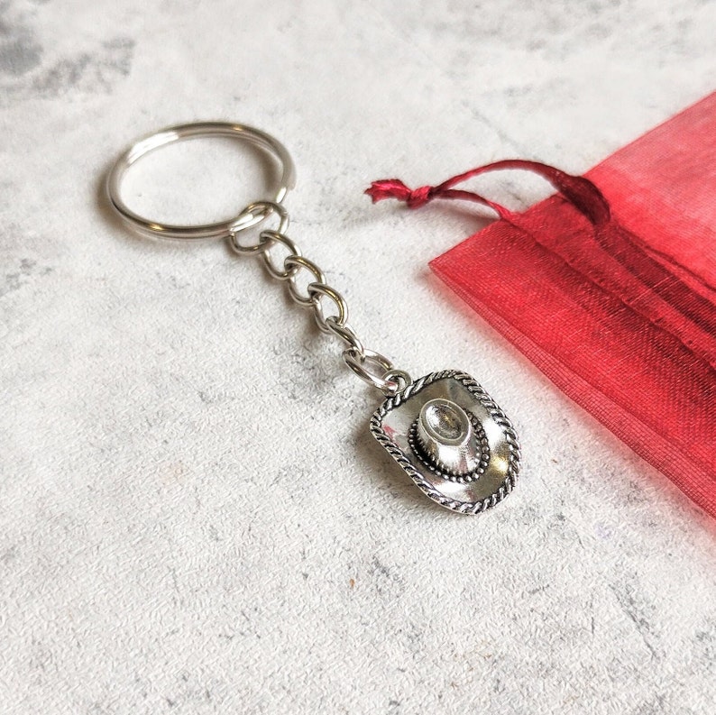 May include: A silver cowboy hat keychain with a silver chain and a silver key ring. The keychain is on a white background.