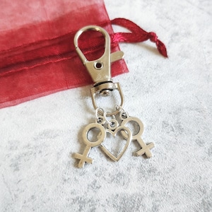 May include: Silver-toned keychain with a lobster clasp. The keychain features three charms: two female gender symbols and a heart. A red organza gift bag is in the background.