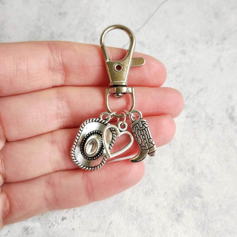 May include: Silver-toned keychain featuring a cowboy hat, heart, and cowboy boot charms. The charms are attached to a metal clasp. The cowboy hat has a detailed brim and center. The boot charm has intricate detailing.