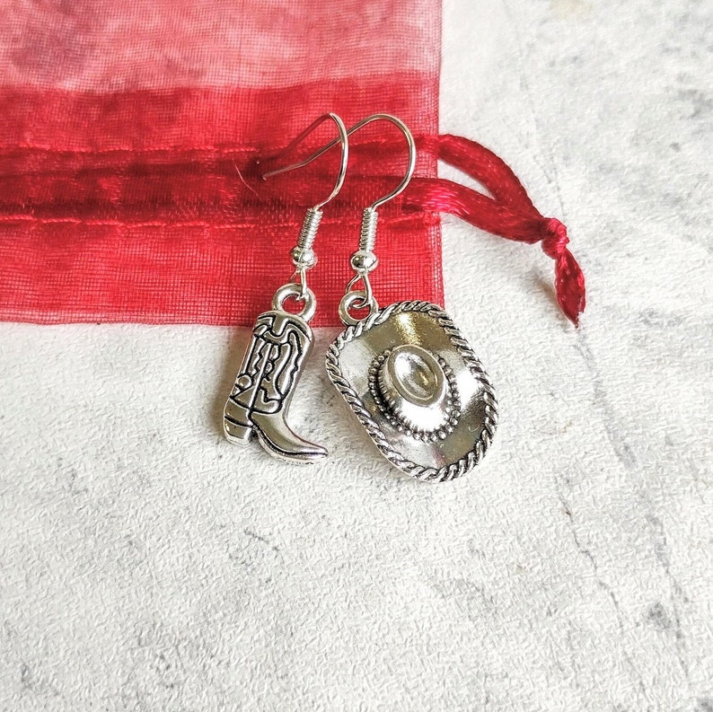 May include: A pair of silver earrings with a cowboy boot and a cowboy hat charm. The earrings are hanging from a red fabric background.