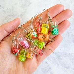 May include: A set of eight colorful fruit-filled jar earrings. The jars are made of clear plastic and filled with colorful fruit slices. The earrings are silver and have a hook closure.