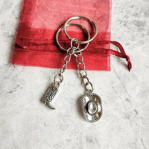 May include: Silver keychain with a cowboy boot and a cowboy hat charm. The keychain is on a red fabric background.