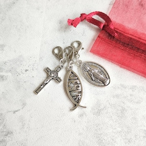 May include: Three silver-toned metal charms with lobster claw clasps. One charm is a crucifix, one is a fish-shaped charm with the word JESUS embossed, and one is an oval medal depicting a figure in robes. A red organza bag is visible.