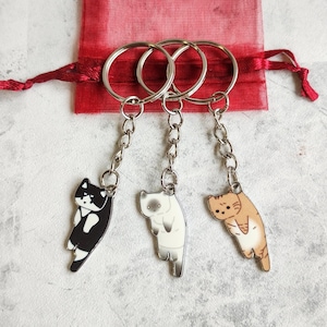 Puede incluir: Tres llaveros con forma de gato con cadenas y anillos plateados. Los gatos son negros y blancos, blancos y grises, y naranjas y blancos. Una bolsa roja con cordón está en el fondo.