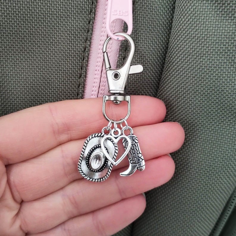 May include: Silver-toned cowboy-themed keychain with a lobster clasp. The charms include a cowboy hat, a heart, and a cowboy boot. The hat has a decorative border and a clear stone in the center. The boot has intricate detailing.