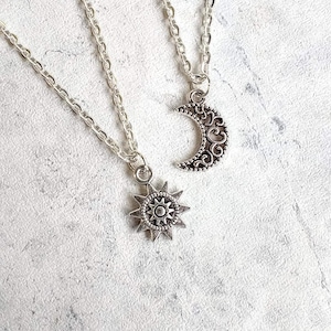 Può includere: Due collane d'argento con un ciondolo a forma di sole e luna. Il ciondolo a forma di sole è un design a stella con un piccolo cerchio al centro. Il ciondolo a forma di luna è una luna crescente con un design decorativo.