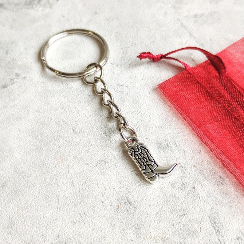 May include: Silver boot keychain with a silver chain and a silver key ring. The keychain is in a red organza bag.