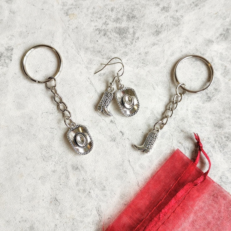 May include: Two silver keychains with cowboy boot and hat charms, and a pair of silver earrings with cowboy boot and hat charms. The keychains and earrings are on a red fabric background.