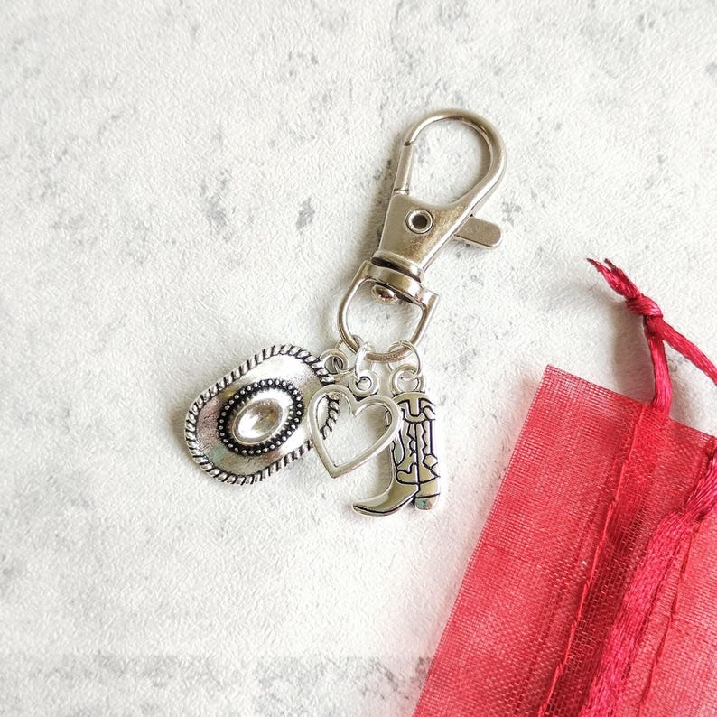 May include: Silver-toned keychain featuring a cowboy hat, heart, and cowboy boot charms. The keychain has a lobster clasp for easy attachment. A small red mesh gift bag is in the background.