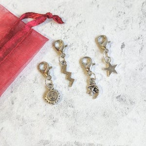 May include: Four silver-toned charms with lobster claw clasps. The charms include a sun with a smiling face, a lightning bolt, a crescent moon with stars, and a five-pointed star. A red fabric gift bag is visible in the background.
