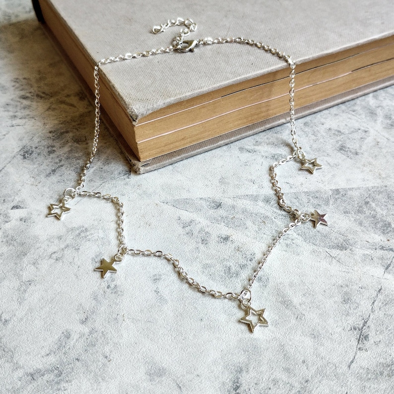 May include: A silver chain necklace with five dangling star charms. The necklace is on a grey textured surface.