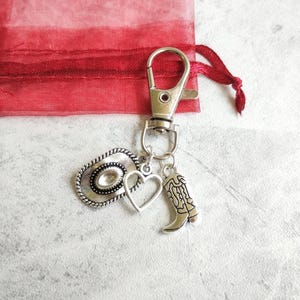 May include: Silver-toned keychain with a cowboy hat, heart, and cowboy boot charms. The keychain is attached to a silver clasp and a red fabric bag with a drawstring.