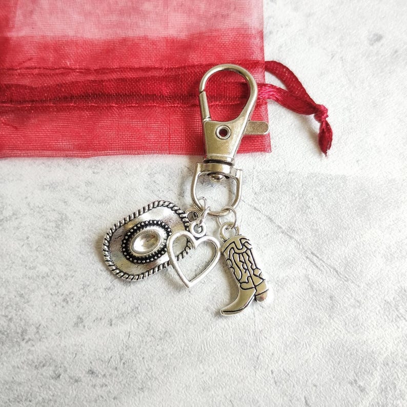 May include: Silver-toned keychain with a cowboy hat, heart, and cowboy boot charms. The keychain is attached to a silver clasp and a red fabric bag with a drawstring.