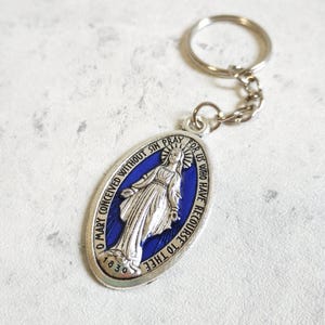 Op de afbeelding: Ovale zilverkleurige sleutelhangerhanger met een blauw middenstuk met een religieuze figuur. De hanger heeft de tekst "O Mary conceived without sin pray for us who have recourse to thee" en het jaartal "1830".