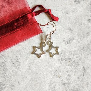 May include: A pair of silver star-shaped earrings with a simple design. The earrings are hanging from a red mesh bag.