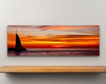 May include: A panoramic canvas print depicting a sunset over the ocean. The sky is ablaze with orange, red, and yellow hues, with a silhouette of a sailboat on the water. The artwork is displayed on a wooden shelf against a white wall.