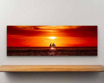 May include: A panoramic canvas print depicting a vibrant sunset over the ocean. The sky is ablaze with shades of red and orange, with a silhouetted sailboat on the water. The artwork is displayed on a wooden shelf against a white wall.