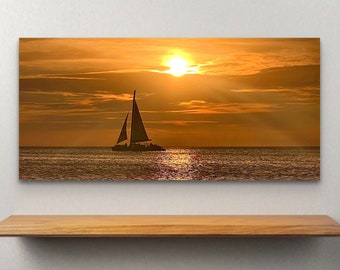 May include: A framed canvas print of a sailboat on the ocean at sunset. The sun is a bright orb in the sky, with rays of light shining through the clouds. The water reflects the golden light.