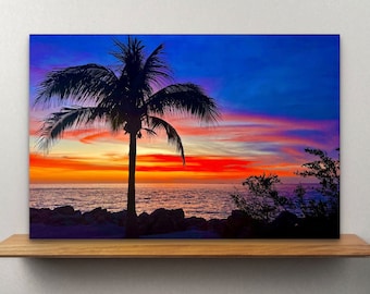 May include: A canvas print of a tropical sunset scene. A silhouetted palm tree stands in front of a vibrant sky with shades of orange, red, and blue. The ocean and rocky shoreline are visible below the horizon.